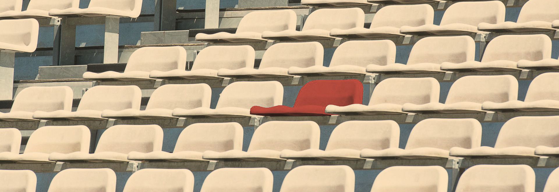 Rows of identical pale stadium seats with one seat in a softened red, standing apart from the rest.