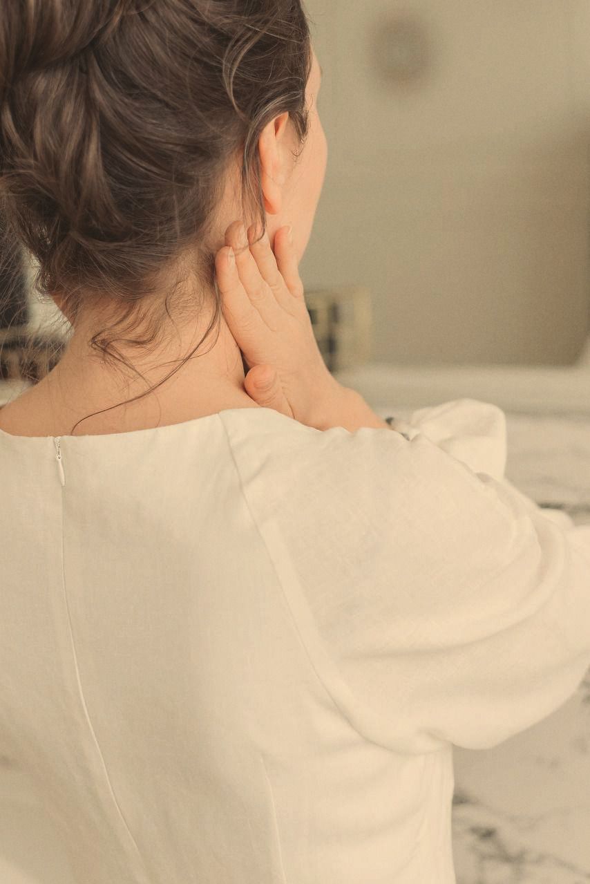 A woman seen from behind, one hand resting at the side of her neck, hair gathered up.