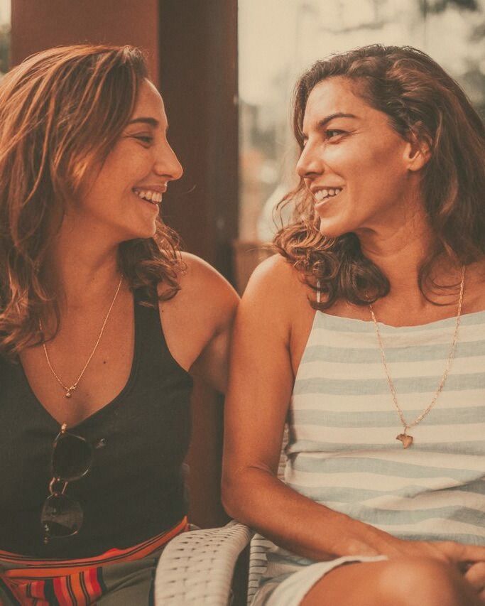 Two women seated close together, laughing mid-conversation in warm evening light.