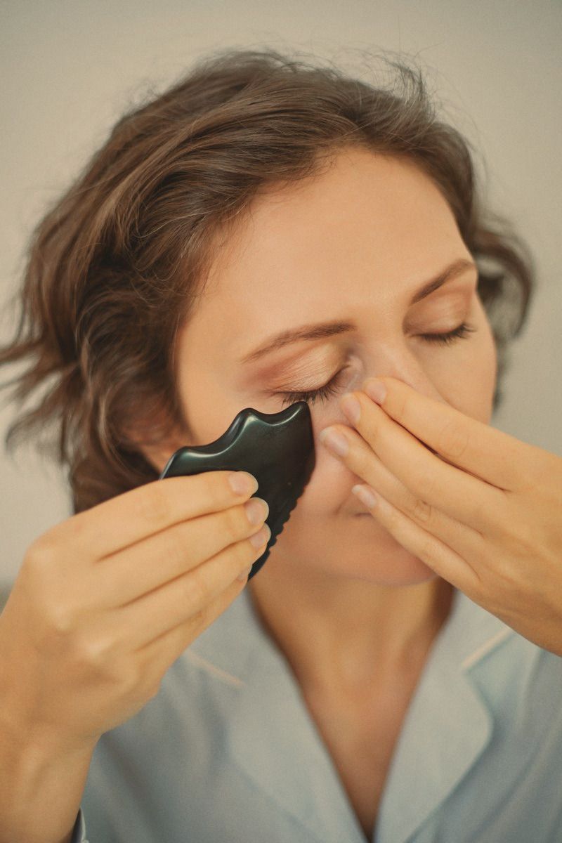 Close crop of a woman's face, eyes closed, working a gua sha stone along her cheekbone.