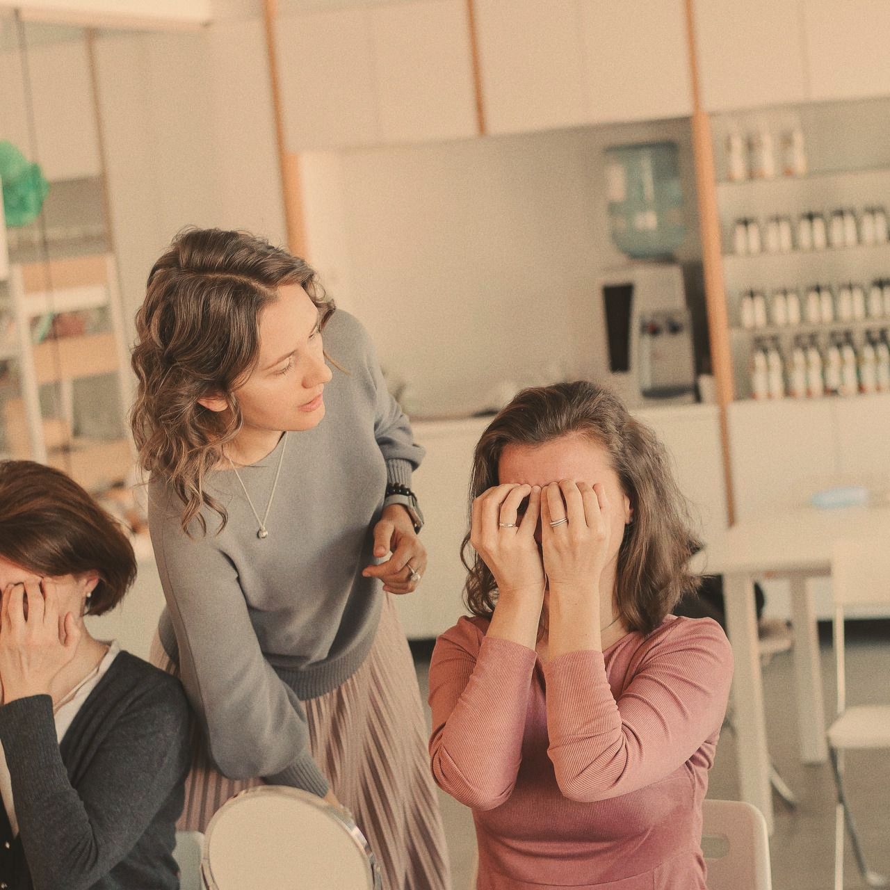 In a bright workshop room, two women sit with their palms over their eyes while a facilitator leans in beside them.