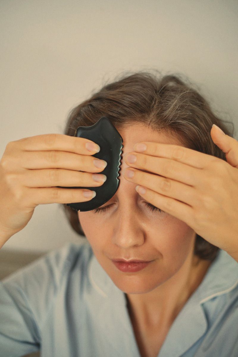 A woman with her eyes closed pressing a black gua sha stone to her forehead with both hands, in a pale blue shirt.