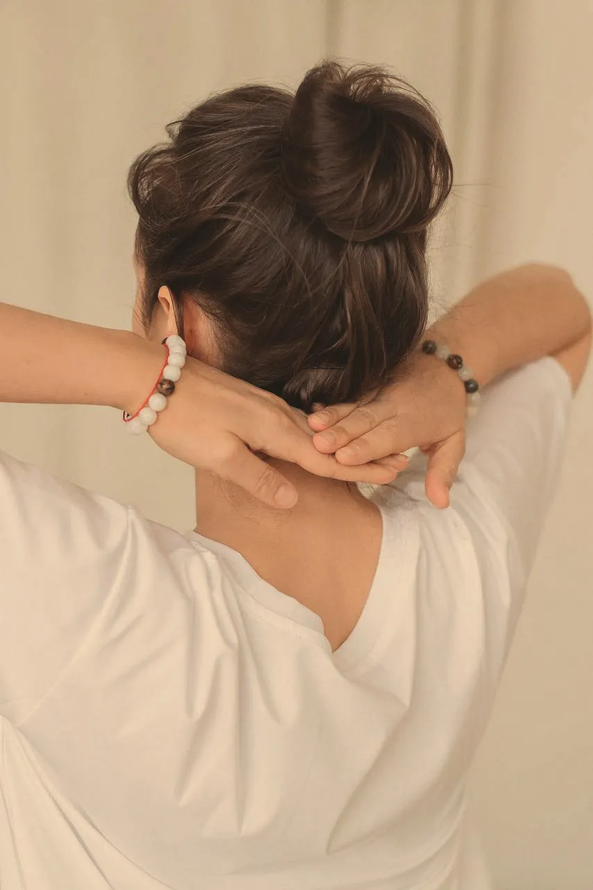 A woman seen from behind, arms raised, gathering her hair into a bun, a white t-shirt and beaded bracelets in soft light.