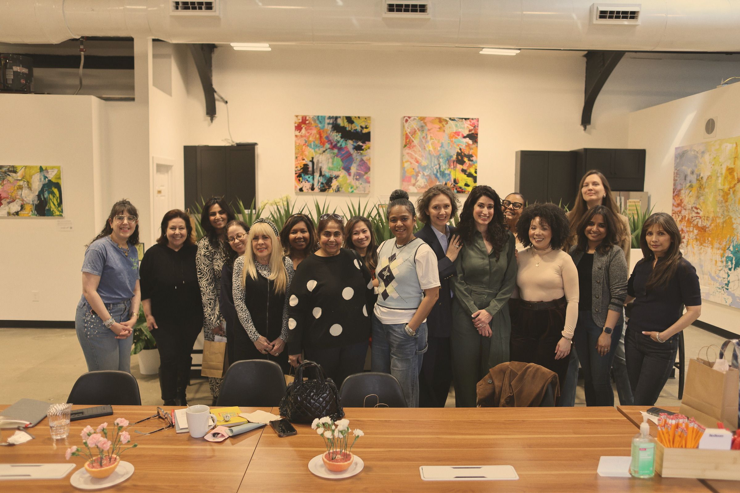 Sixteen women standing together in a bright loft space hung with colourful abstract paintings, a long wooden table in front of them.
