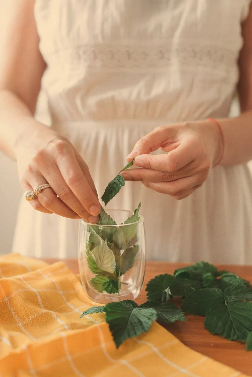 Hands in a white dress dropping fresh green leaves into a glass on a wooden table with a yellow checked cloth.