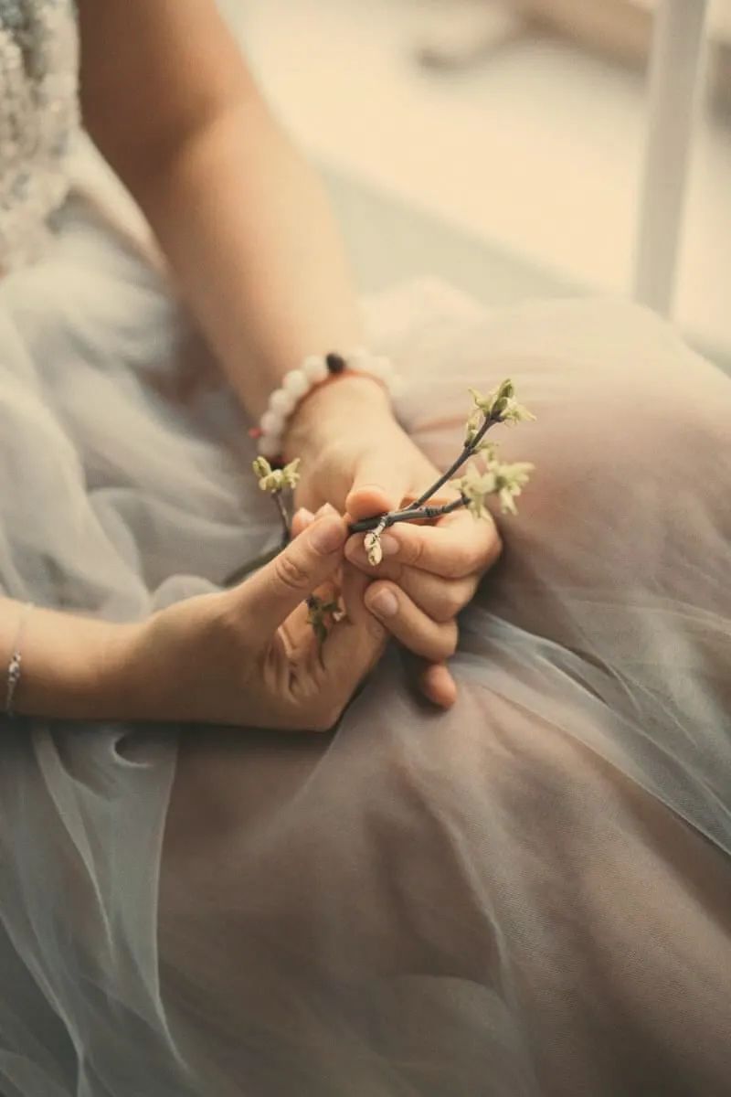 Hands resting in a lap of pale tulle, holding a small flowering twig, a beaded bracelet on one wrist.