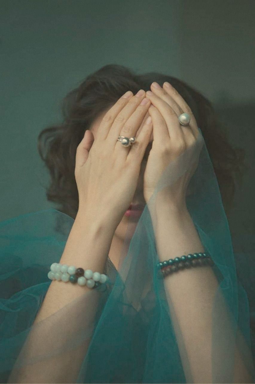 A woman covering her face with both hands behind a veil of teal tulle, rings and beaded bracelets catching the light.