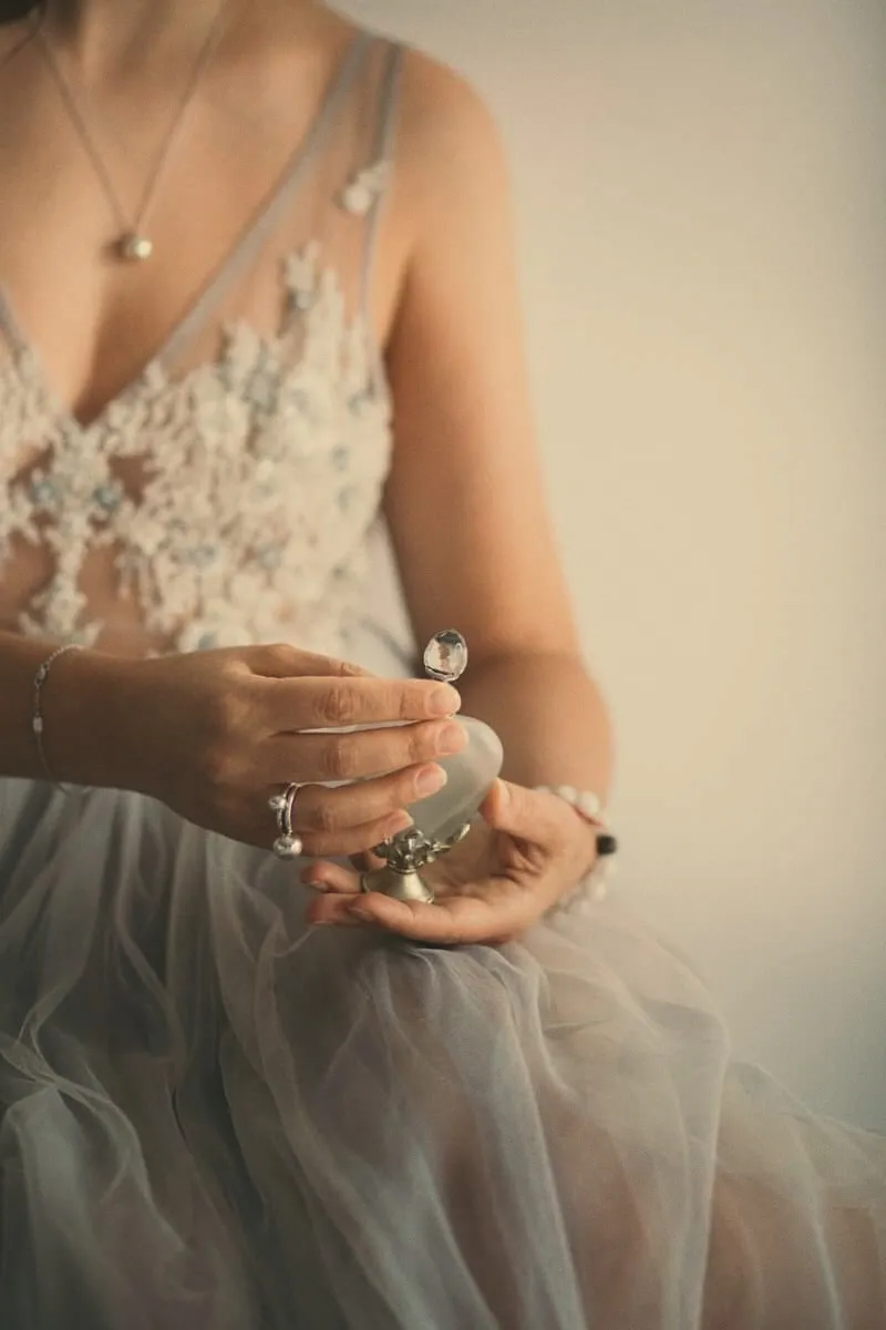 Hands in a lap of grey tulle holding a small crystal perfume bottle, rings and a bracelet catching the light.