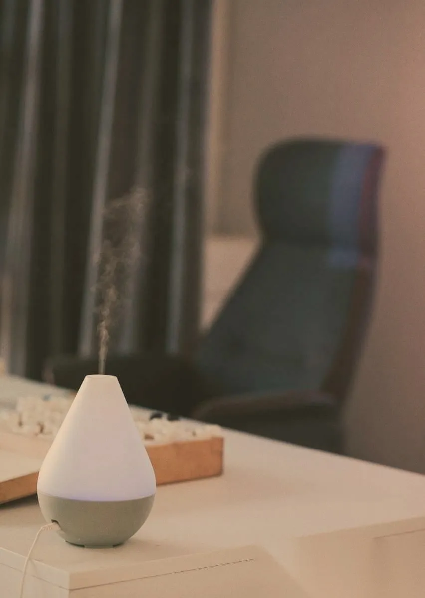 A diffuser releasing a thin column of mist on a desk, a wooden tray of stones beside it and a dark office chair behind.