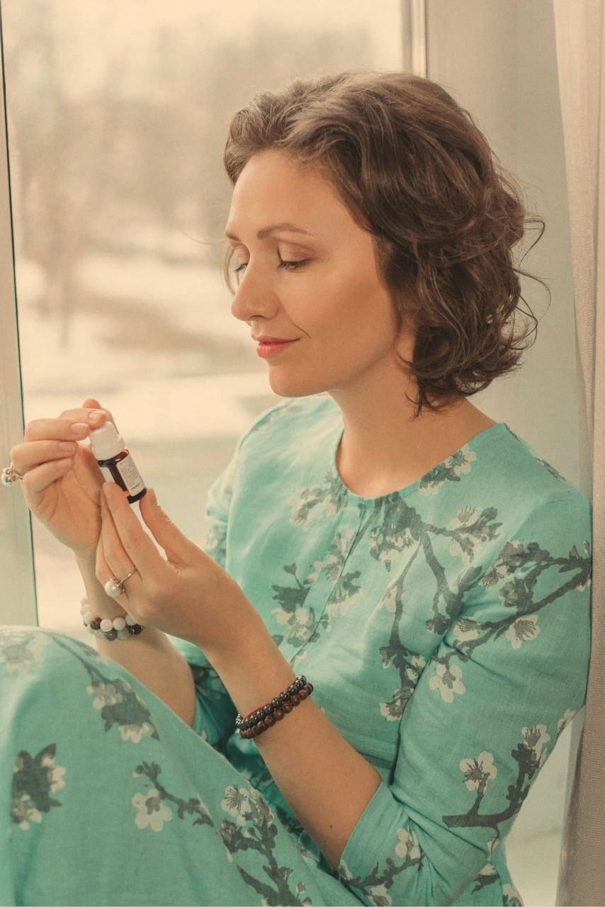 A woman in a teal floral dress seated by a window, turning a small amber bottle in her hands, snow-covered trees outside.