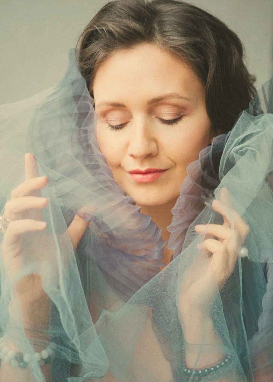 A woman with her eyes closed, framed by sheer fabric she holds up near her jaw with both hands.