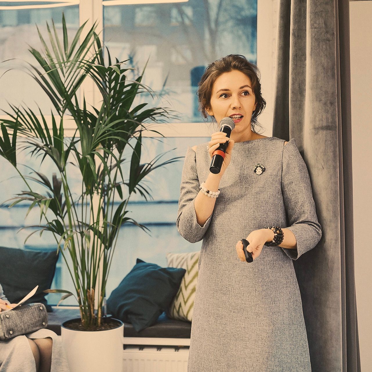 Natalia speaking into a handheld microphone in a daylit room beside a large plant.
