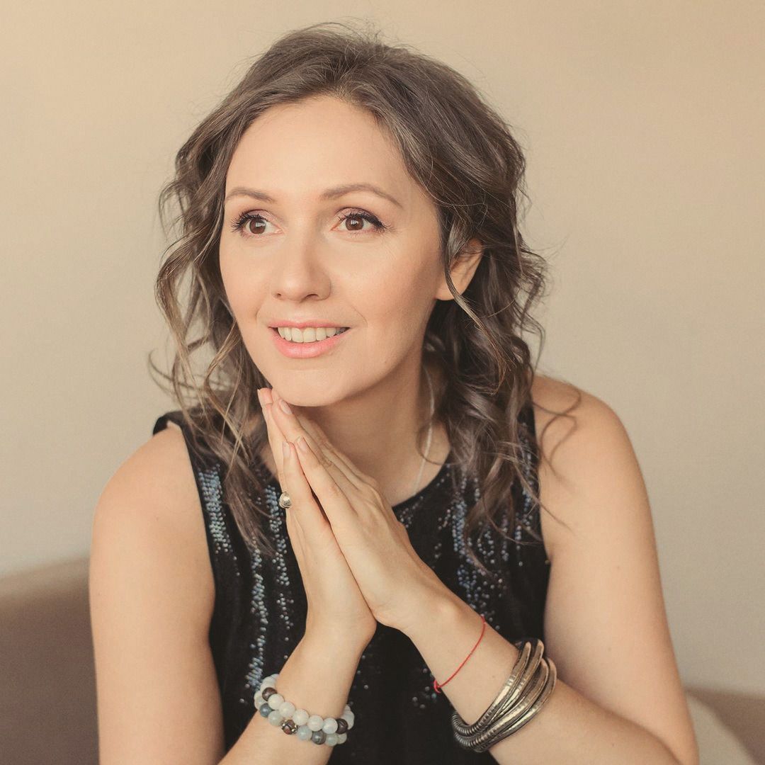 A woman with curly hair, hands clasped at her throat, smiling and looking to the side.