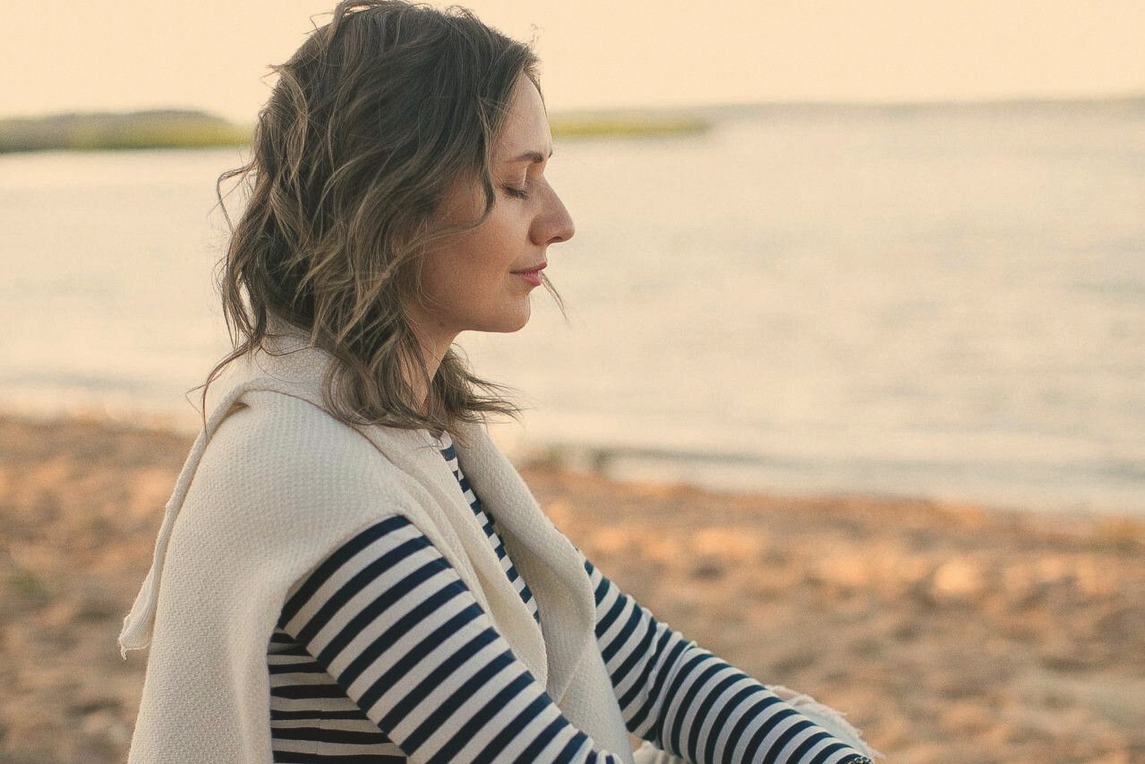 A woman sits cross-legged on the sand by the water, eyes closed, hands resting together.