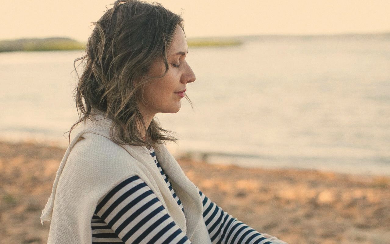 A woman sits by the water in evening light, eyes closed, face turned toward the sea.