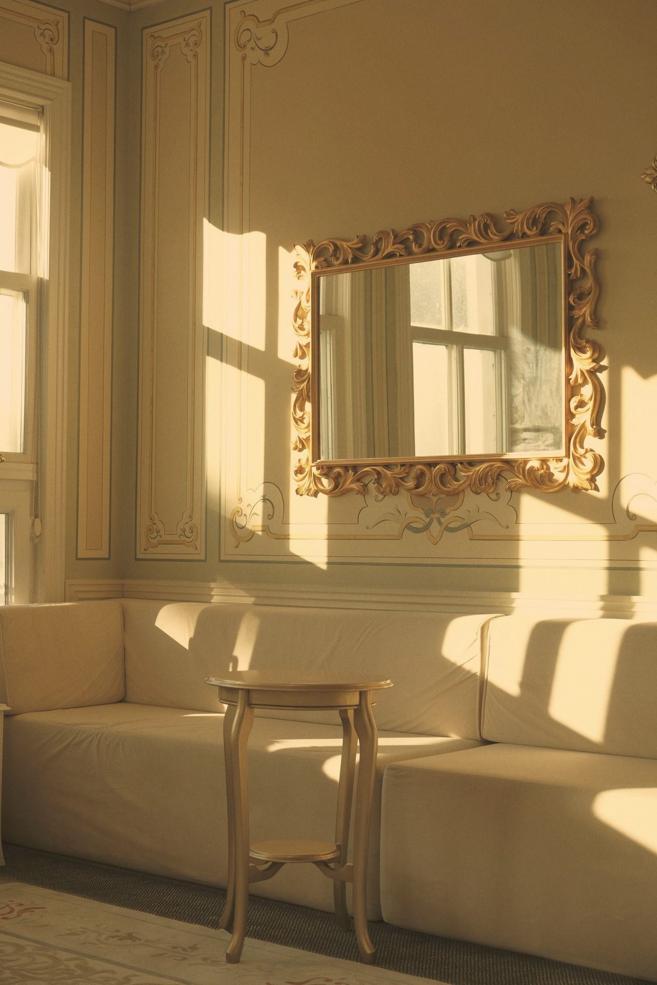 A gilded mirror on a sunlit plaster wall above a pale sofa, nobody in frame.