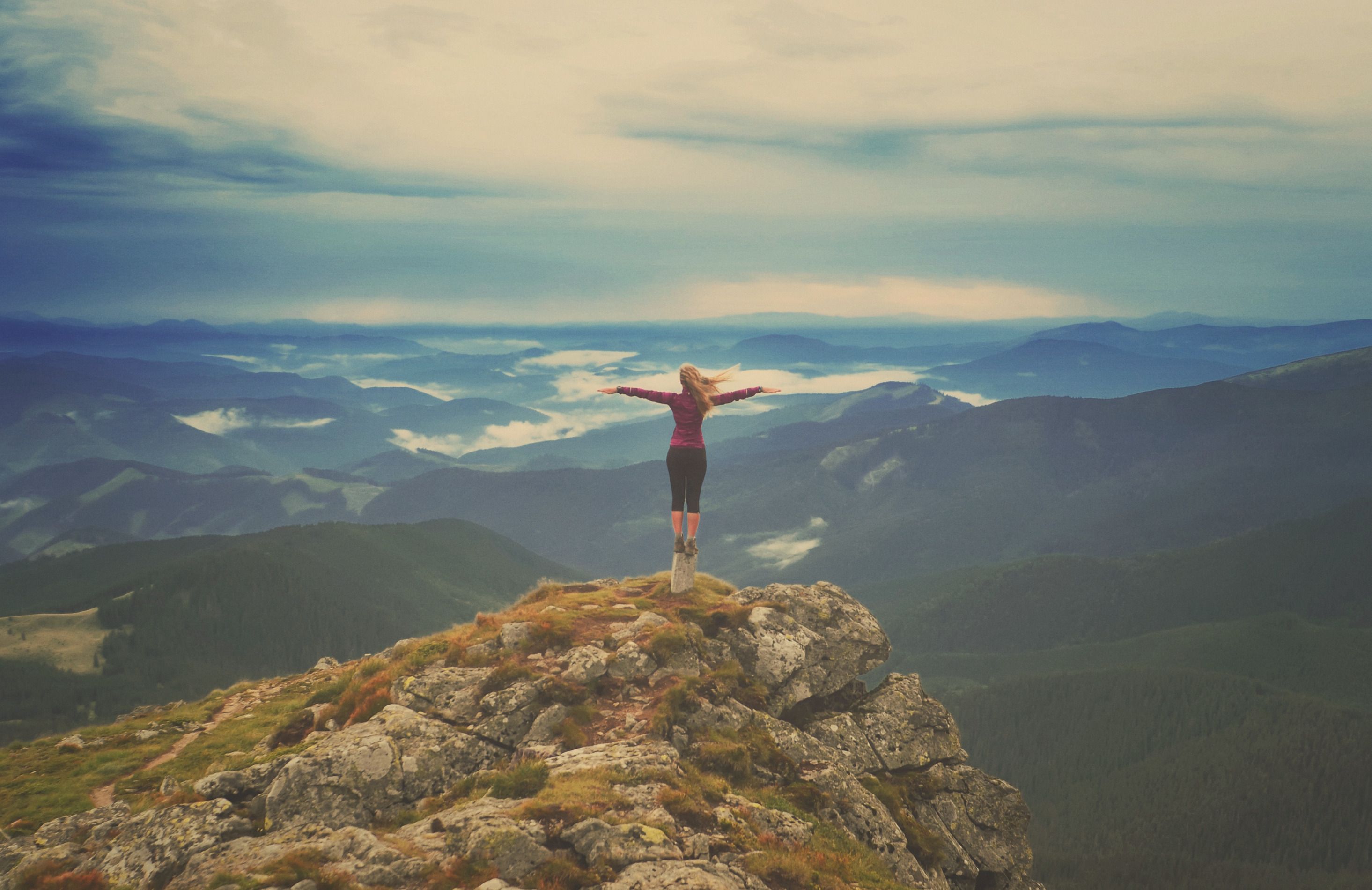 A woman standing on a mountain summit, arms outstretched, layered peaks and clouds behind her.
