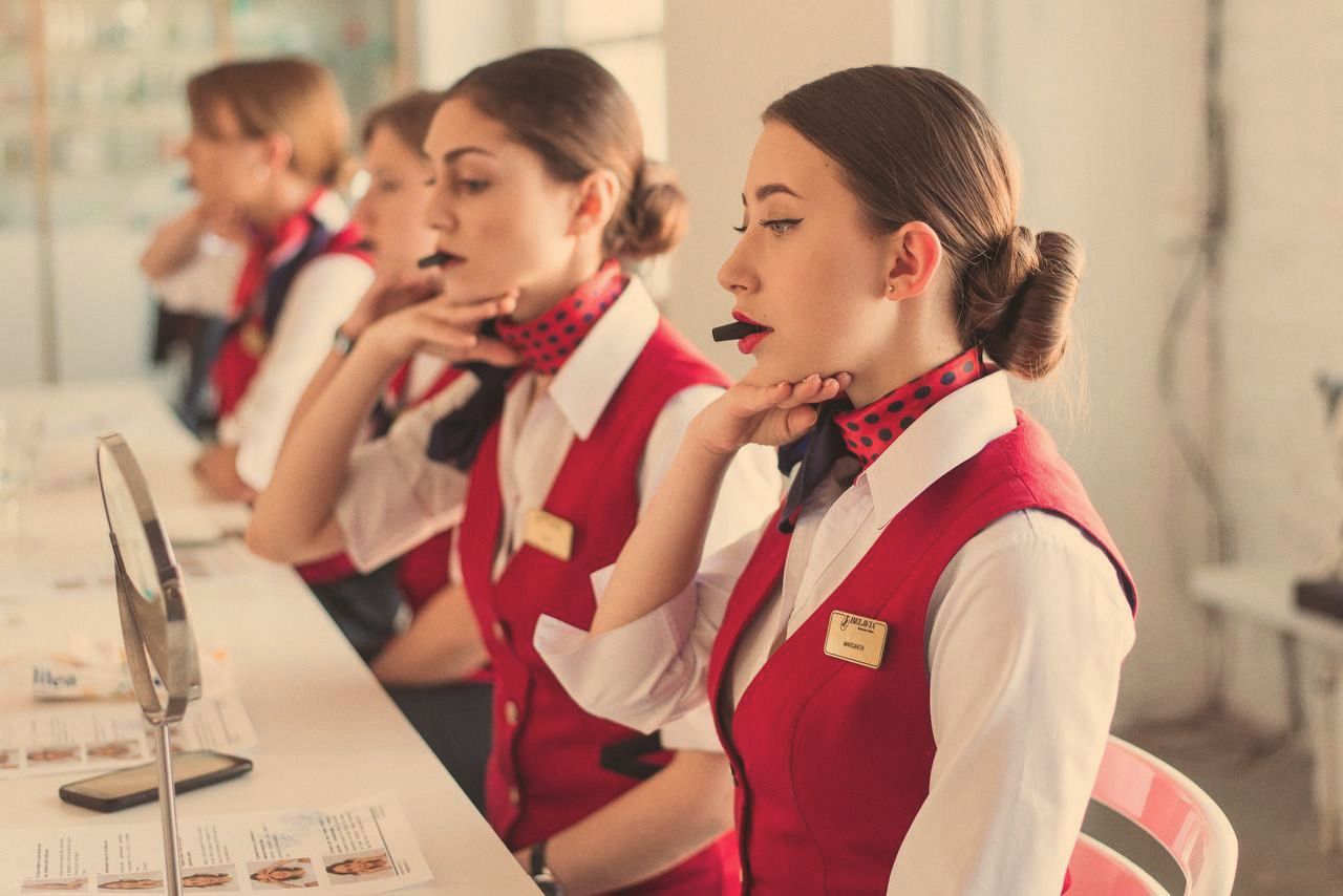Flight attendants in red uniforms and polka-dot scarves sit in a row at a long table, each studying a small mirror during a grooming session.