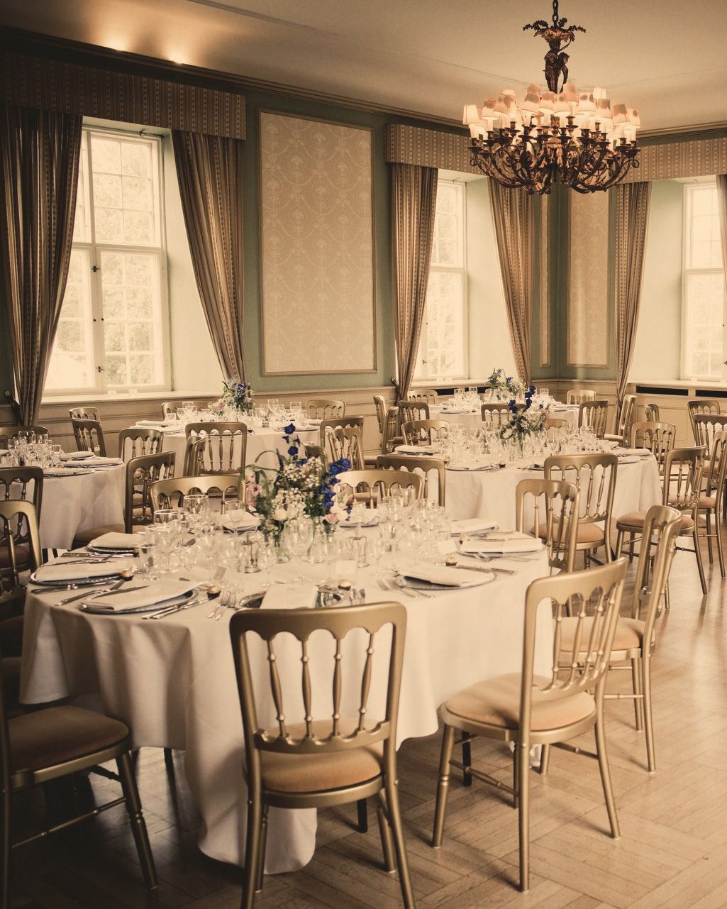 A formal event dining room with round tables set in white linen and gold chairs beneath a chandelier.