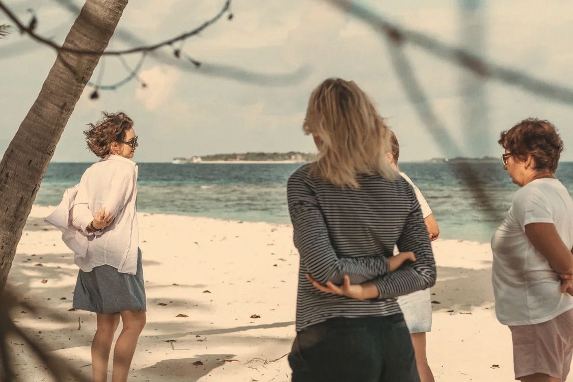 Four women stand together on a white-sand beach, turquoise water and a small island behind them, listening to a woman in a white shirt.