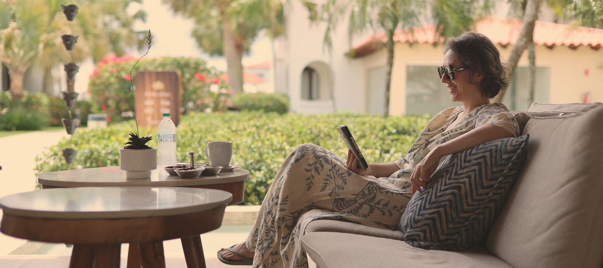 A woman relaxing on a resort patio in sunglasses, reading a tablet.