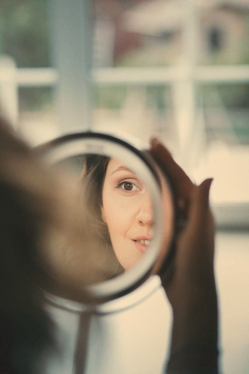 A woman's eye reflected in a round hand mirror she holds up, morning light and a window blurred behind her.