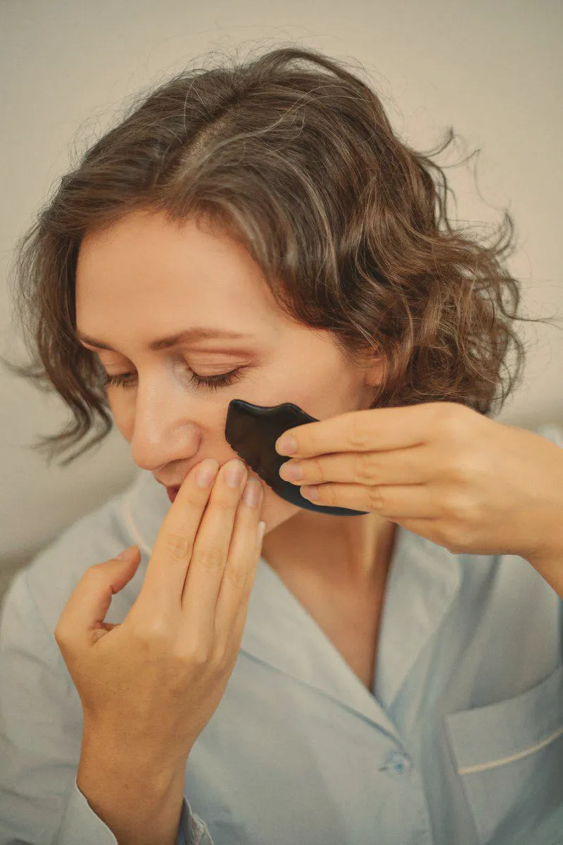 Close crop of a woman's face, eyes closed, drawing a gua sha stone along her cheekbone.