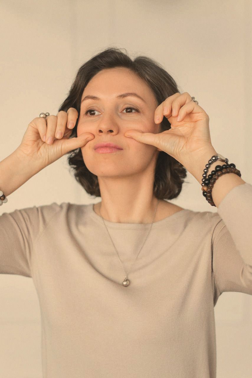 A woman facing the camera, fingertips pressed to her cheeks mid facial practice, against a plain wall.