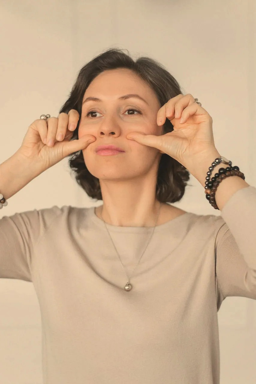 A woman in a beige top gently pinching both cheeks between her fingers, eyes turned to the side, in even studio light.