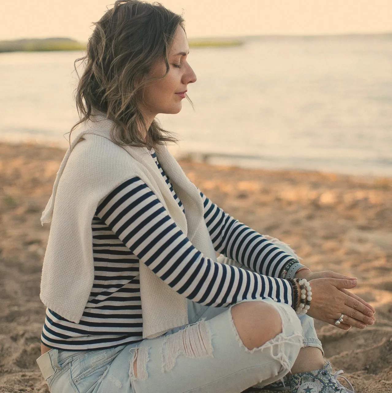 A woman sitting cross-legged on a sandy shore at sunset, eyes closed, hands resting loosely in her lap.
