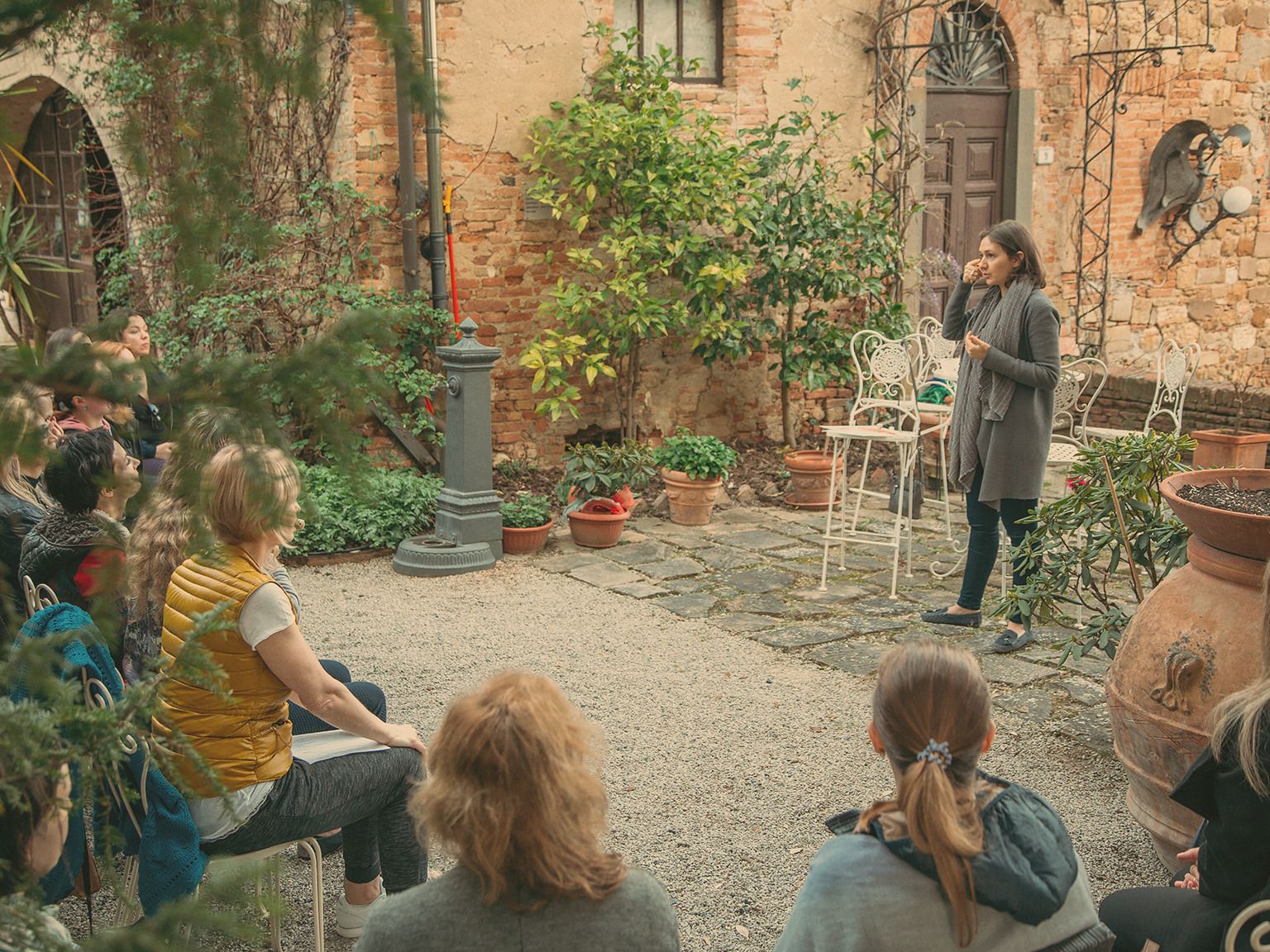 A woman speaking to a small group of women seated in a walled stone courtyard planted with potted greenery.