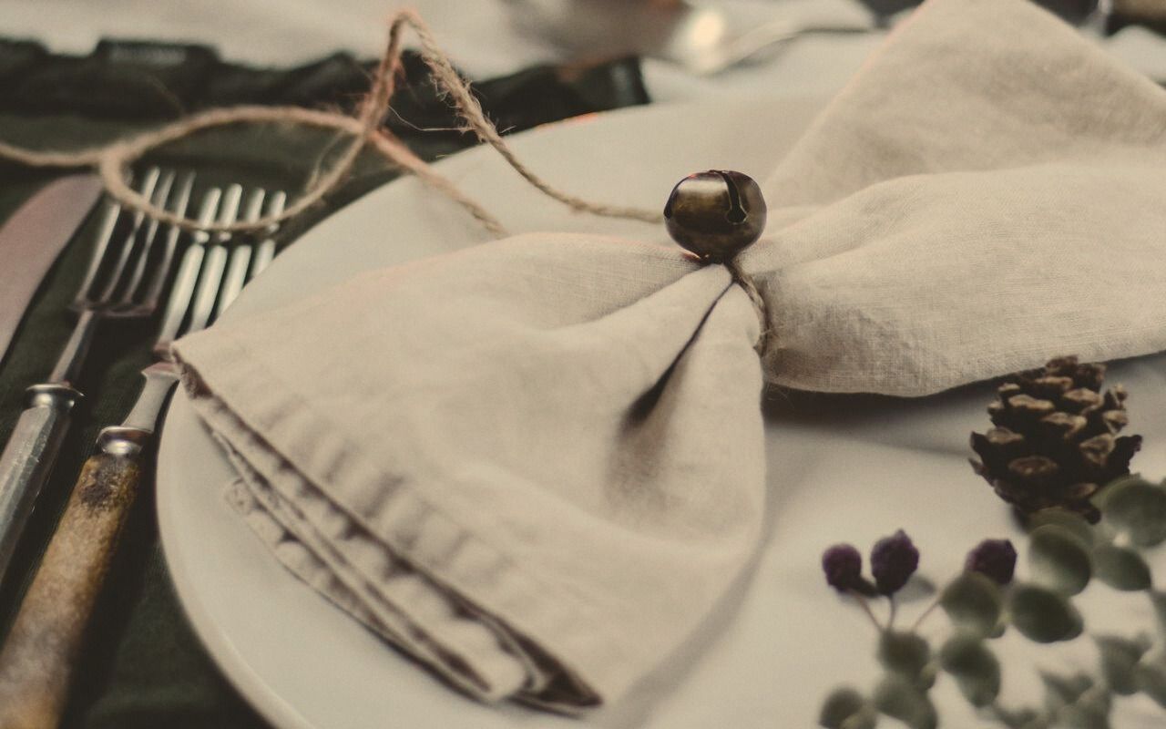 A folded linen napkin tied with twine on a white plate, beside worn cutlery, a pinecone and a sprig of eucalyptus.